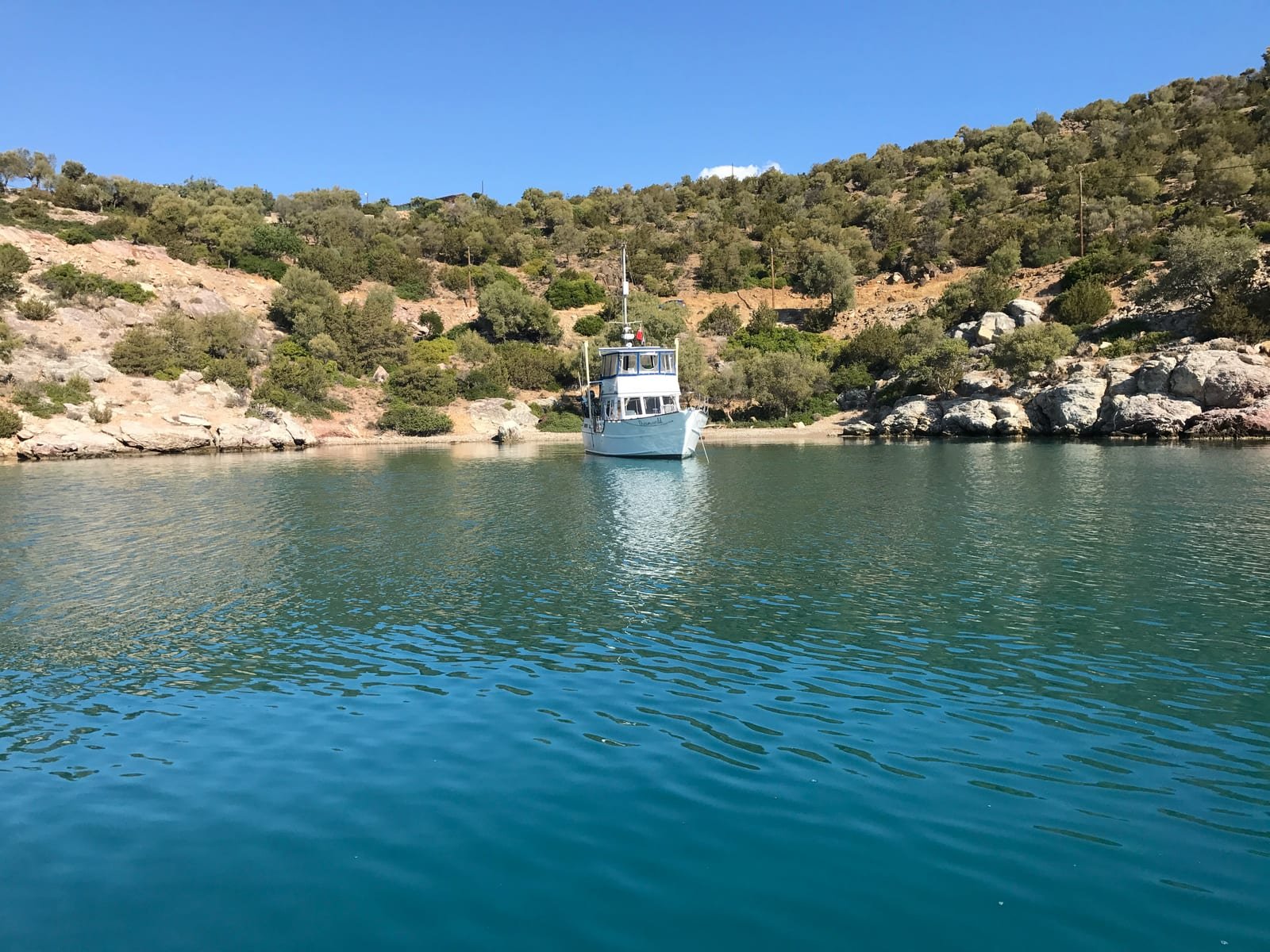 Dreamworld lying at anchor in a still turquoise cove in Greece, close under a rocky hillside covered in olive trees.