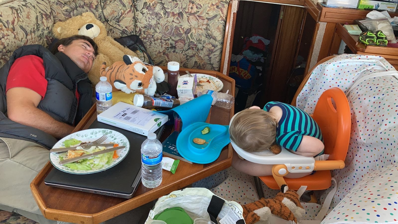 The galley table aboard Dreamworld after a meal: Simon asleep on the bench among the plates with a teddy bear and a stuffed tiger beside him, and the baby asleep face-down on the tray of his highchair.