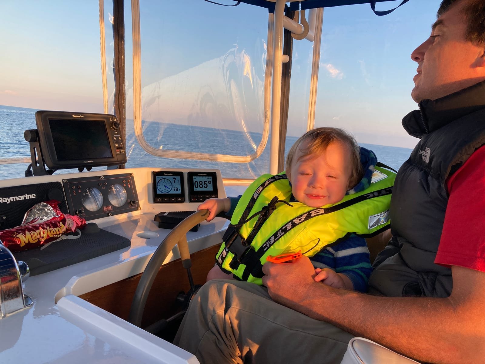Simon at Dreamworld’s wheel at sunset, his small son on his lap in a yellow lifejacket with one hand on the helm. The instruments read a heading of 085 degrees and flat sea runs to the horizon.