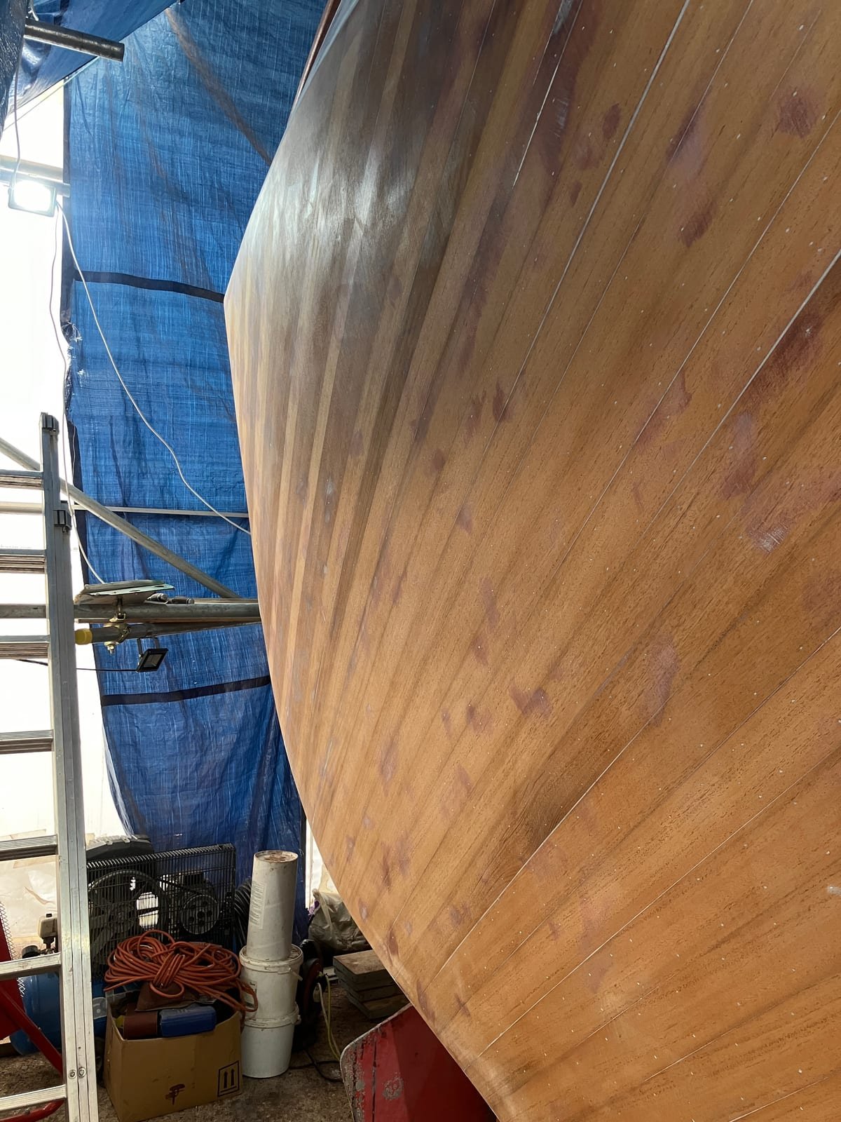 The bare planked mahogany hull of Dreamworld curving away from the bow, sanded back to the wood during her refit, under a blue tarpaulin shelter.